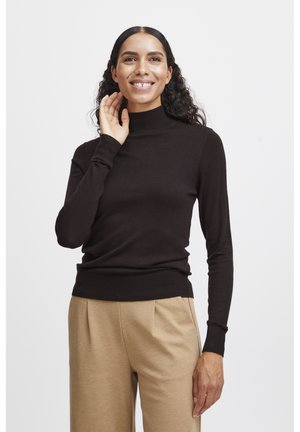 Pull à col roulé brun foncé, design ajusté, poignets et ourlet côtelés, associé à un pantalon beige, porté par une femme souriante.