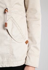 Veste beige clair en toile avec une poche avant, un cordon de serrage avec des accents en cuir et un ourlet cousu. Le matériau a une texture lisse.