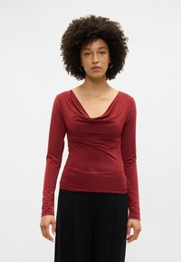 Dolga majica s fitting krojem v temno rdeči barvi, z drapiranim izrezom in gladkim, raztegljivim materialom. Kombinirana z ohlapnimi črnimi hlačami.