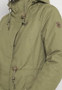 Chaqueta con forro interior de color verde oliva, con capucha, botones, cordones, un parche con logo y una tela texturizada.