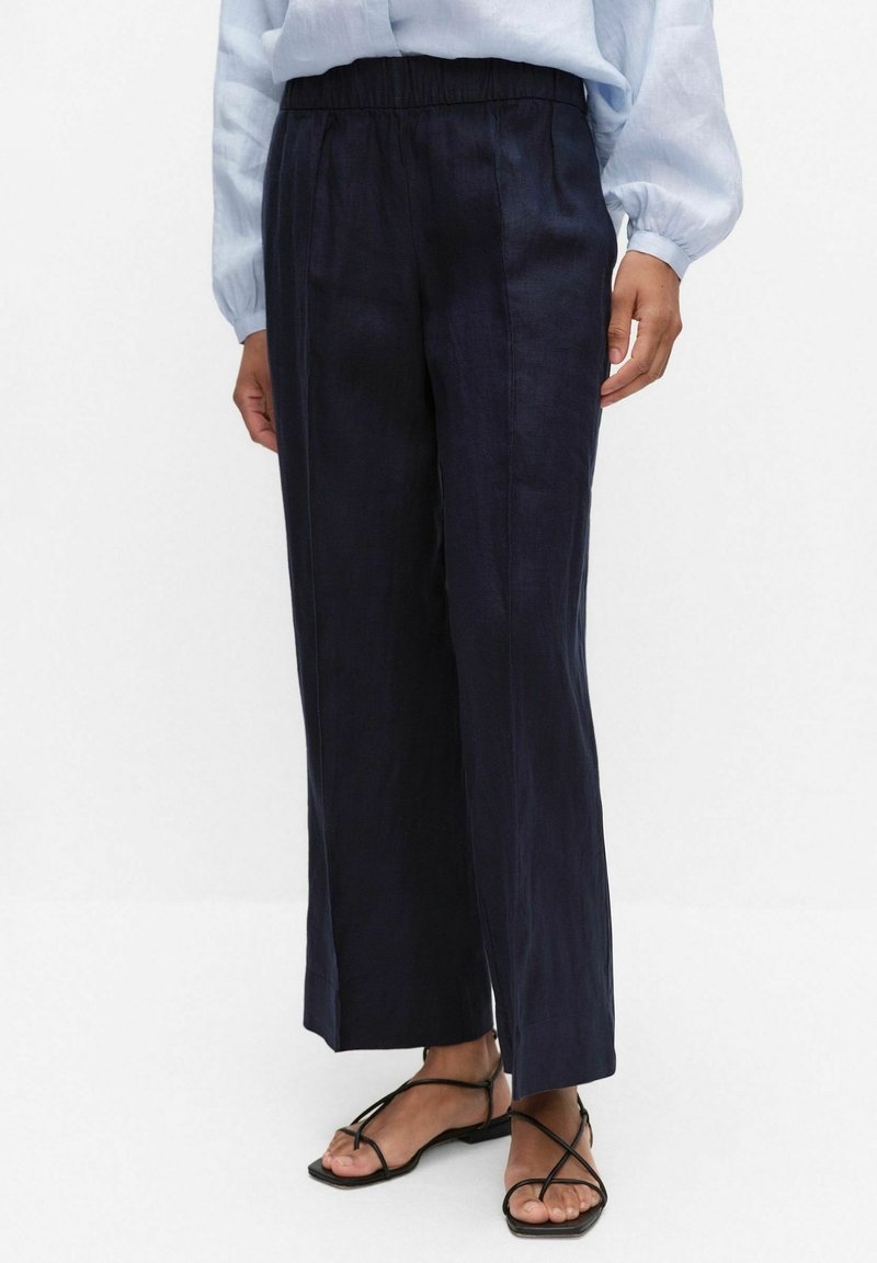 Persoon draagt marineblauwe wijde broek met elastische taille en zwarte sandalen met bandjes en platte zool, gecombineerd met een lichtblauwe blouse met lange mouwen.