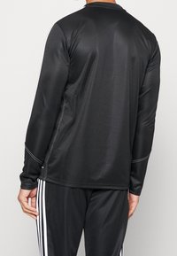 Musta pitkähihainen urheilupaita, joka on tehty sileästä, kevyestä kankaasta. Paidassa on valkoinen tikkaus ja pyöreä pääntie, ja se on yhdistetty vastaaviin housuihin.