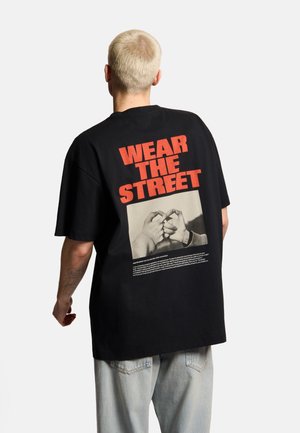 Osoba z krótkimi blond włosami, ubrana w czarny oversize T-shirt z czerwonym napisem "WEAR THE STREET" i zdjęciem gestu ręką na plecach.