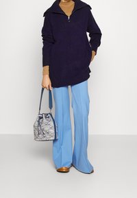 Navy knit sweater with a high collar, paired with light blue trousers. The model holds a textured bucket bag featuring a subtle pattern and blue accents.