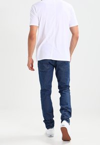 Vit kortärmad t-shirt i kombination med blå denimjeans. T-shirten har en central söm; jeansen har en klassisk passform med bakfickor.