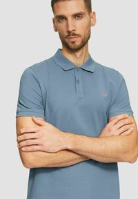 Helles blaues Poloshirt aus Baumwolle, mit klassischem Kragen, einer Zweiknopf-Leiste, kurzen Ä sleeves und einem kleinen gestickten Logo auf der Brust.