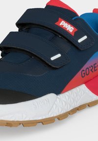 Gros plan d'une basket bleu marine et rouge avec deux bandes Velcro, semelle blanche portant les mentions "STORM," et "GORE" imprimées sur le côté.
