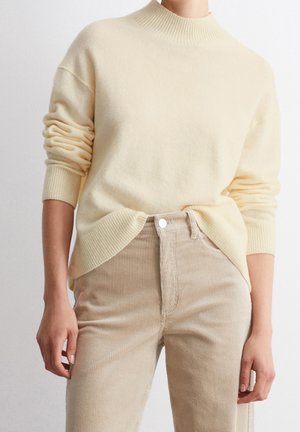 Person iført en cremefarvet ribstrikket sweater med ærmerne opsmøgede og beige manchestrebukser, hænderne hvilende ved siden, foran en ensfarvet baggrund.