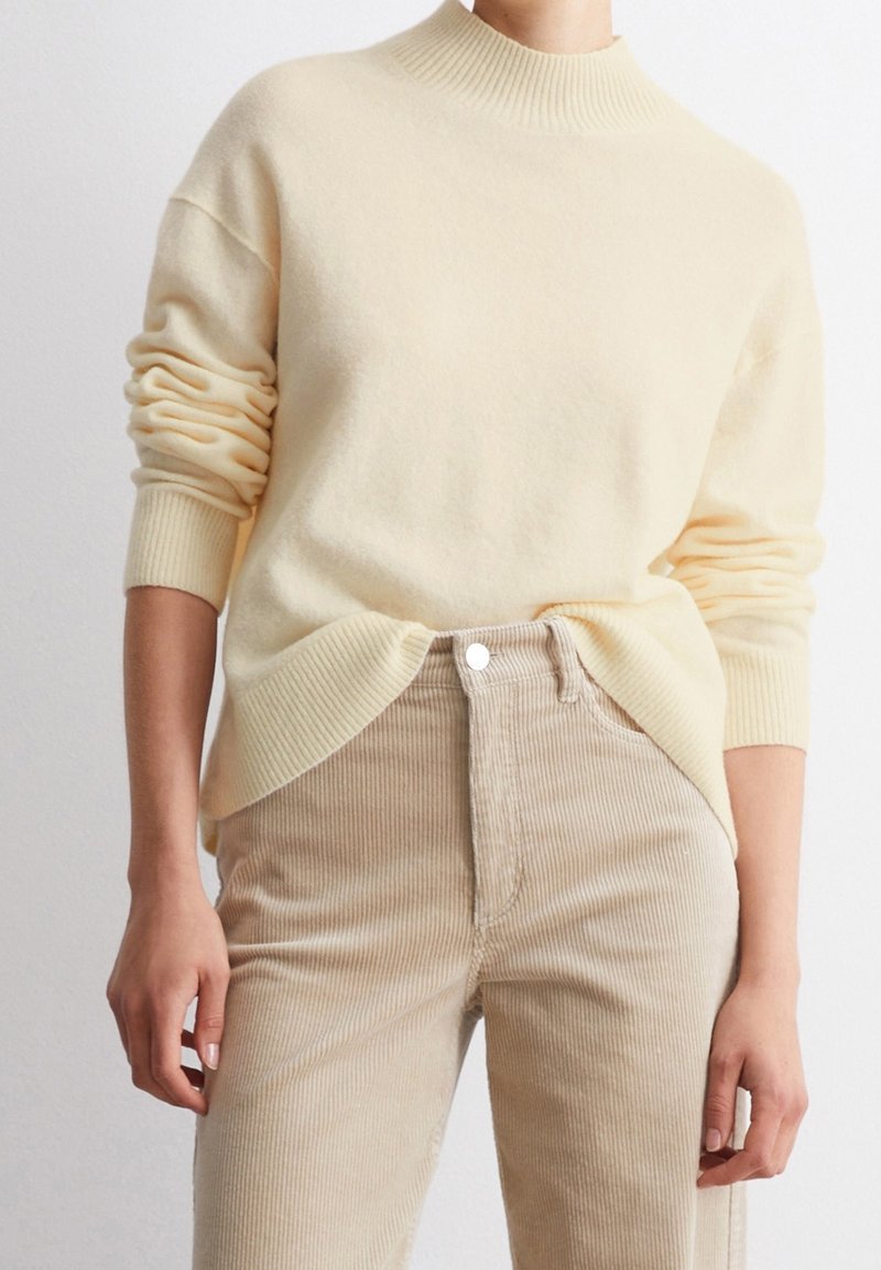 Personne portant un pull côtelé crème avec les manches remontées et un pantalon beige en velours côtelé, mains reposant le long du corps, sur un fond uni.