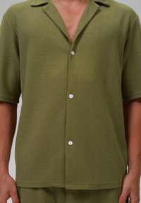 Cămașă țesută, cu mânecă scurtă, de culoare verde măsliniu, cu un model texturat, guler cu decupaj și nasturi albi pe față.