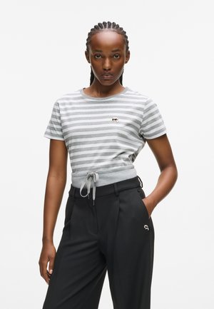 Woman with braided hair wearing a gray and white striped t-shirt and high-waisted black trousers with hands in pockets on a white background.