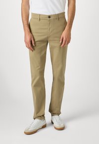 Chinos de algodón color caqui con un corte slim, bolsillos frontales y un cierre de botón único, combinados con zapatillas blancas y una camisa blanca lisa.