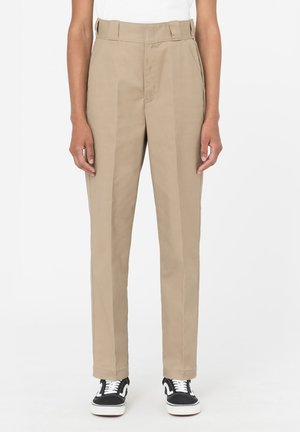 Beige højtaljede bukser med lige ben, der har frontfolder og bæltestropper. Stoffet har en glat tekstur. Kombineret med sorte sneakers.