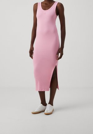 Femme portant une robe midi rose sans manches avec une fente sur le côté et des baskets blanches, debout devant un fond gris uni.