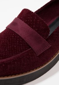 Chaussure slip-on bourgogne avec un dessus texturé, présentant une large bande et un bord festonné, ainsi qu'une semelle noire plus épaisse pour la stabilité.