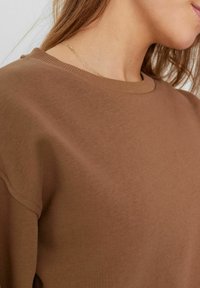 Barna pamut pulóver kerek nyakkivágással és bordás szegéllyel, sima textúrával és laza szabással. Diszkrét varrási részlet az alján.