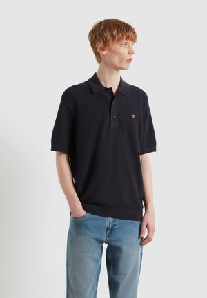 Giovane uomo che indossa una polo nera a maniche corte con trama e jeans azzurri, in piedi con una mano nella tasca, guardando di lato.