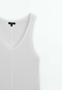 Débardeur blanc en coton avec un col en V profond, texture côtelée et couture contrastante. Présente une couture centrale à l'avant et une étiquette au niveau du cou.