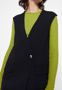 Svart ärmlös väst med V-ringning, med en enda knapp och två framfickor, lager på en ljusgrön långärmad skjorta.