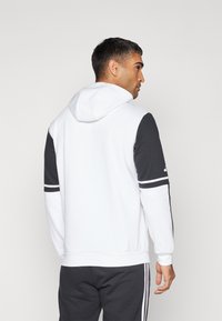 Hoodie mit einem weißen Körper, dunkelgrauen Ärmeln und weißen Streifenakzenten. Hergestellt aus einem weichen, strukturierten Stoff für zusätzlichen Komfort.