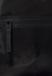Čierna vodeodolná taška s hladkým povrchom, vybavená výrazným zipsom, detailom loga a minimalistickým dizajnom.