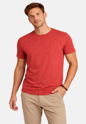 Hombre de pie con la mano en el bolsillo de unos pantalones beige, vistiendo una camiseta roja de manga corta con un pequeño logo en el pecho.