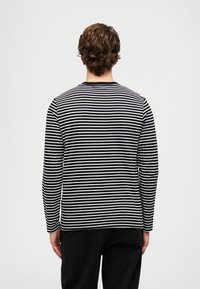 Egy férfi háttal áll, fekete és fehér vízszintes csíkos hosszúujjú pólót és fekete nadrágot visel, egy sima fehér háttér előtt.