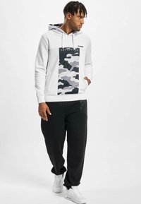 Hvit hettegenser med grå camo-panel, med et logo og lommer. Kombinert med svarte bukser og hvite sneakers, noe som skaper et avslappet antrekk.
