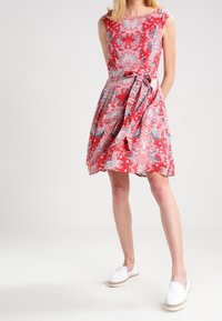 Rotes Blumenkleid mit tailliertem Oberteil, schwingendem Rock und seitlichem Bindeband. Baumwollstoff mit feinen Mustern; kombiniert mit weißen Espadrilles.