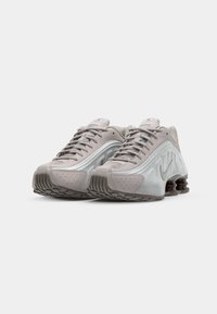 Des baskets Nike gris clair présentent un design épuré avec des panneaux texturés et un accent argenté brillant. La semelle comprend des éléments noirs élastiques pour un meilleur amorti.
