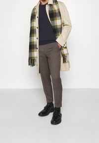 Man i beige kappa, svart tröja, grå byxor, svarta monk strap-skor och en fransad rutiga scarf i gult, vitt och svart.