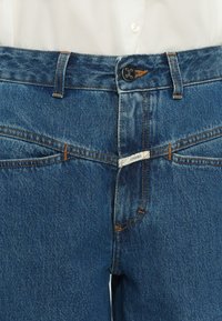 Gros plan sur un jean en denim bleu moyen avec des coutures orange, un bouton métallique marqué "CLOSED" et une étiquette blanche avec le texte "CLOSED" à la taille.