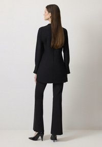 Blazer negro ajustado con mangas largas, combinado con pantalones de campana negros y tacones de punta, visto desde la parte de atrás, textura suave.