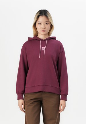Sweat à capuche bordeaux avec poche kangourou, capuche à cordon et une petite étiquette de logo. Manches longues avec poignets côtelés, associé à un pantalon marron.