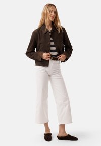 Braune Wildleder-Cropped-Jacke mit Spitzkragen, Knopfleiste und Brusttasche, kombiniert mit weißen weit geschnittenen Hosen und dunklen Slippern.