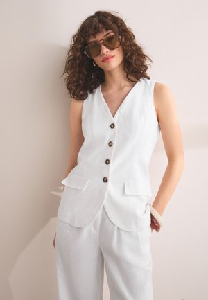 Donna con capelli ricci che indossa grandi occhiali da sole marroni, gilet bianco senza maniche con bottoni, pantaloni bianchi coordinati e un braccialetto leggero, che posa all'interno.