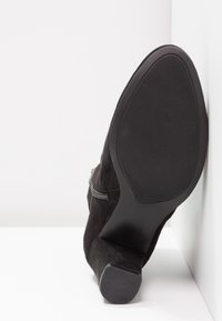 Botte cheville en daim noir avec une semelle extérieure texturée, un détail de fermeture éclair latérale et un talon effilé. Fini lisse et mat sans motifs visibles.