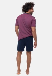 Homme avec des cheveux bouclés portant un t-shirt rayé bordeaux et un short marine, vu de dos, debout pieds nus contre un fond blanc.