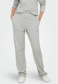 Persoon in lichtgrijze sweatpants met handen in de zakken en witte sneakers, staande tegen een effen achtergrond.