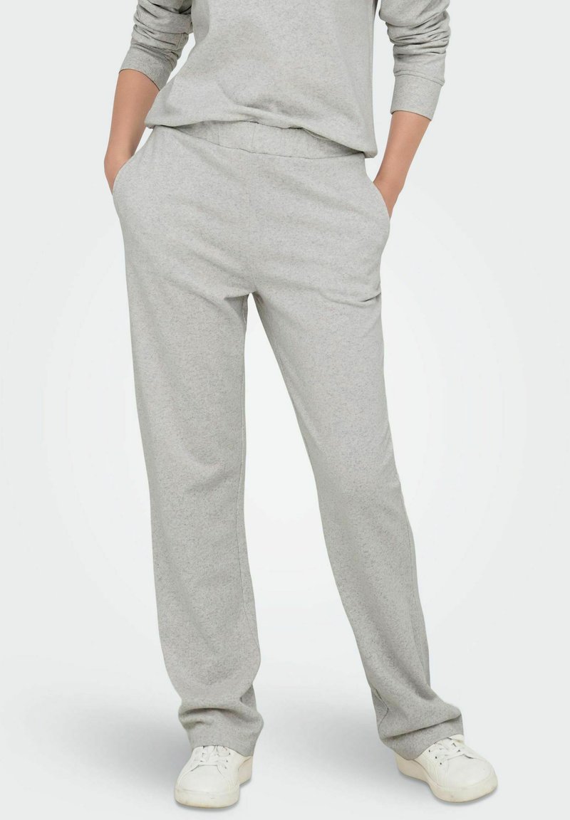 Persoon in lichtgrijze sweatpants met handen in de zakken en witte sneakers, staande tegen een effen achtergrond.