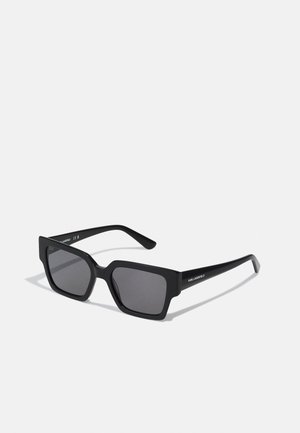 Occhiali da sole neri con finitura opaca, lenti grigie e stanghette eleganti con il marchio sul lato. Design semplice e geometrico.