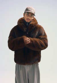 Brun pälsjacka med hög krage och dragkedja, med en plyschig textur. Bärs över grå lösa byxor, med en keps i rutigt mönster.