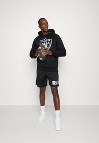 Sudadera negra con el logo de "Raiders", pantalones cortos negros con la letra "RAIDERS", zapatillas blancas, sosteniendo un balón de rugby con un patrón. Fondo neutro.