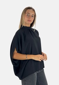 Schwarzes, locker sitzendes Oberteil mit hohem Kragen und kurzen Ärmeln, aus einem strukturierten Stoff gefertigt, kombiniert mit dunklen Jeans.