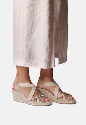 Nohy v béžových klínových sandálech s květinovou výšivkou a překříženými pásky, doplněné světle zbarvenou sukní do kotníků.