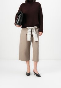 Maglione borgogna lavorato a maglia, pantaloni beige alla caviglia con cravatta a righe bianche in vita e zoccoli neri con tacco. Pochette nera in mano.