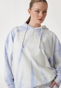 Svijetloplavi i bijeli tie-dye dukserica s labavim krojem, s prednjim logotipom i kapuljačom s vezicama. Mekana tkanina s glatkom teksturom.