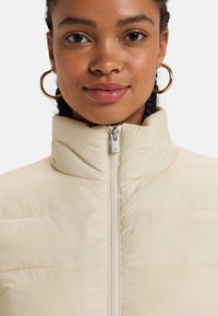 Pufferjacka i ljus krämfärg med hög krage, framf zipper och horisontell quiltning. Har en slät, glänsande tygtextur.