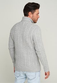 Grau gestrickter Pullover mit hohem Kragen, gerippten Bündchen und Saum. Die Textur zeigt komplizierte geflochtene Muster und vermittelt ein gemütliches Erscheinungsbild.