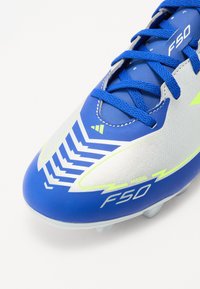 Chaussure de football bleue au design épuré, avec des accents argentés, des motifs zigzag proéminents et des lacets bleus. Équipée d'une semelle à crampons.